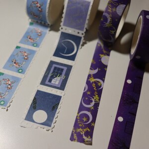 May include: Four rolls of decorative washi tape with different designs. The first roll has a blue background with white koi fish. The second roll has a blue background with a crescent moon, a window, and a white circle. The third roll has a purple background with gold crescent moons, stars, and the text "dreams are free". The fourth roll has a purple background with gold trees and white dots.
