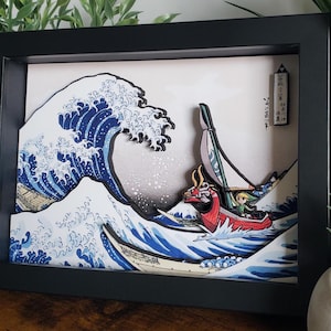 May include: A layered shadow box art piece in a black frame featuring a stylized blue and white ocean wave inspired by The Great Wave off Kanagawa, with a red dragon-headed boat carrying a small green-clad figure sailing through the water.