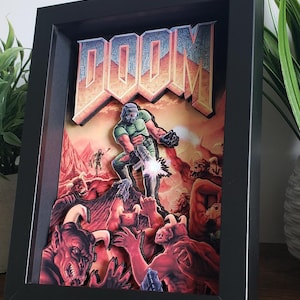 May include: A 3D shadow box art piece featuring the DOOM logo at the top and a central figure in green armor firing a weapon at various red, horned demon creatures. The scene is set against a fiery orange background, all enclosed in a black frame.