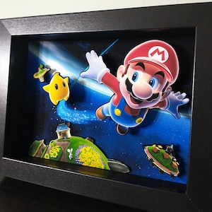 May include: A three-dimensional shadow box art piece featuring Mario floating in space with a yellow star character. The scene includes a small green planet with a house and a floating island against a dark blue starry background, all encased in a black frame.