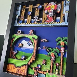 May include: A 3D diorama of a level from the video game Sonic the Hedgehog. The diorama features a blue sky, green grass, palm trees, and a loop-de-loop. There are several characters from the game, including Sonic, Tails, and Knuckles. The diorama is framed in black.