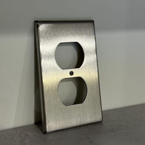 May include: A brushed metal electrical outlet cover with two oval openings and a small circular opening. The cover is rectangular with a slightly textured surface and a metallic sheen. The item is positioned against a light gray wall.