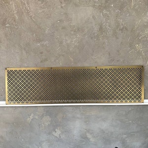 Può includere: Una griglia di ventilazione in metallo dorato con un motivo decorativo ripetitivo. La griglia di ventilazione è rettangolare e ha una finitura liscia e lucida.