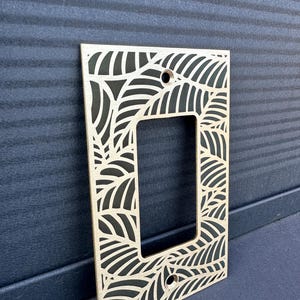 May include: A wooden light switch plate with a laser-cut leaf design. The plate has a rectangular opening for the switch and two screw holes. The leaf pattern is cut out, revealing the dark surface behind it. The plate is made of light-colored wood.