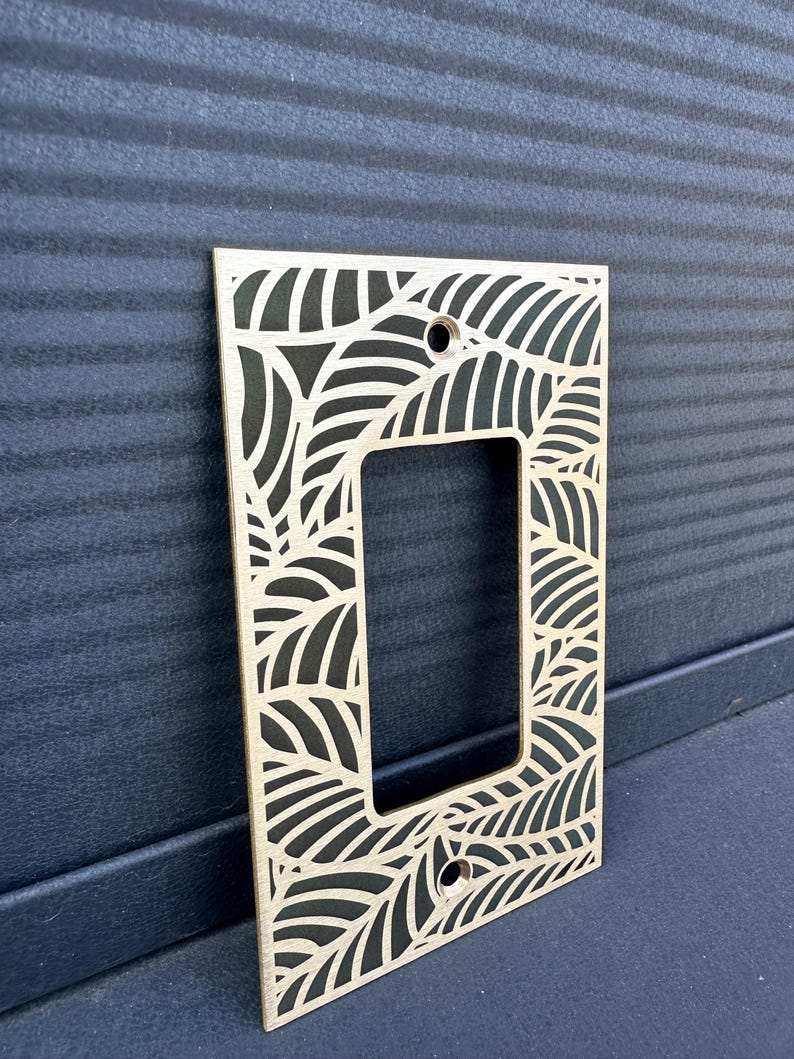 May include: A wooden light switch plate with a laser-cut leaf design. The plate has a rectangular opening for the switch and two screw holes. The leaf pattern is cut out, revealing the dark surface behind it. The plate is made of light-colored wood.