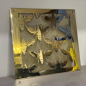 Può includere: Un'opera d'arte da parete quadrata in metallo color oro con un motivo ripetuto di figure stilizzate di falene o farfalle. Gli insetti sono ritagliati dal metallo, rivelando uno sfondo a rete. L'opera d'arte ha una superficie lucida e riflettente.