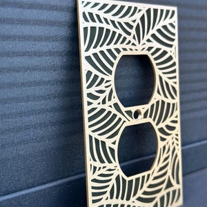 May include: A decorative light wood electrical outlet cover with a leaf design. The cover has two circular openings for the outlets and is surrounded by a laser-cut pattern of leaves. The background is a dark blue.