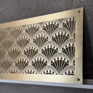 May include: A rectangular, Art Deco-style brass vent cover with a repeating fan-shaped pattern. The cover has a dark mesh backing and is mounted on a white surface against a textured gray wall. The brass has a brushed finish.
