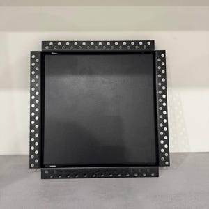 Könnte beinhalten: Ein quadratisches, schwarzes Metallobjekt mit einem perforierten Rahmen. Der Rahmen hat gleichmäßig verteilte Löcher um den Umfang. Das Objekt scheint eine Montagehalterung oder eine ähnliche Strukturkomponente zu sein.