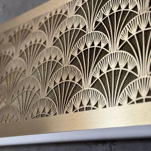 May include: A rectangular, Art Deco-style metal panel with a repeating fan-shaped design. The panel is a warm gold color, with a dark mesh backing visible through the cut-out pattern. The bottom edge is white, and the panel is mounted on a gray surface.