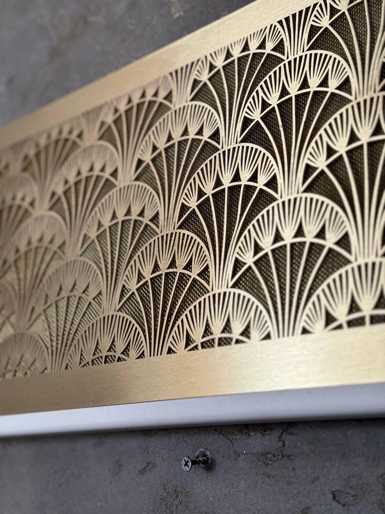 May include: A rectangular, Art Deco-style metal panel with a repeating fan-shaped design. The panel is a warm gold color, with a dark mesh backing visible through the cut-out pattern. The bottom edge is white, and the panel is mounted on a gray surface.