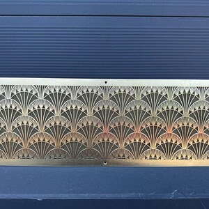May include: A rectangular, gold-colored metal decorative panel with an Art Deco fan pattern. The panel has a series of repeating fan-shaped designs, with small holes at each corner and along the bottom edge.