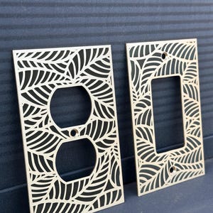 May include: Two wooden light switch covers with a leaf design. One cover has a round outlet opening, and the other has a rectangular switch opening. The covers are made of light-colored wood and have a leaf pattern cut out.