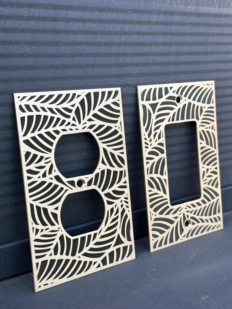 May include: Two wooden light switch covers with a leaf design. One cover has a round outlet opening, and the other has a rectangular switch opening. The covers are made of light-colored wood and have a leaf pattern cut out.