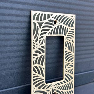 May include: A light-colored wooden switch plate cover with a leaf design. The rectangular cover has a cutout for the switch and two screw holes. The leaf pattern is cut out of the wood, creating a decorative border.