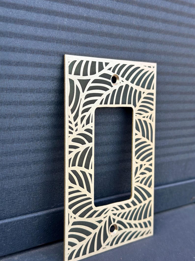 May include: A light-colored wooden switch plate cover with a leaf design. The rectangular cover has a cutout for the switch and two screw holes. The leaf pattern is cut out of the wood, creating a decorative border.