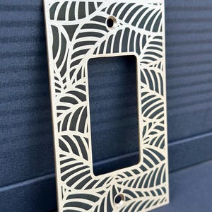 May include: A decorative light switch cover made of light-colored wood. The cover features a rectangular cutout for the switch and is intricately laser-cut with a leaf pattern. The design is set against a dark background.