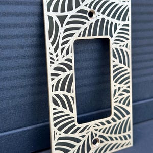 May include: A rectangular light wood switch plate cover with a leaf design cut out. The cover has a rectangular opening in the center and two holes for screws. The leaf pattern is repeated around the edges of the cover.