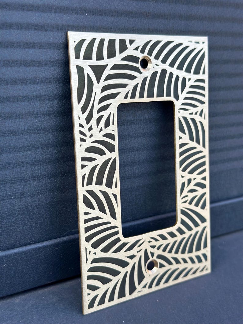 May include: A rectangular light wood switch plate cover with a leaf design cut out. The cover has a rectangular opening in the center and two holes for screws. The leaf pattern is repeated around the edges of the cover.