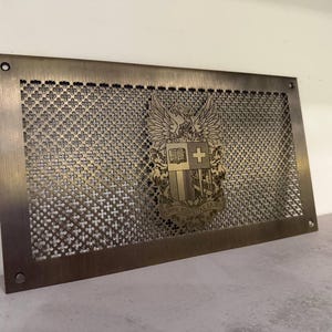 Peut inclure: Une grille de ventilation rectangulaire en métal avec un motif décoratif de petites croix et un emblème détaillé. L'emblème présente un bouclier, une croix, un livre et un aigle avec l'inscription "AMOR FIDELITAS LAUDIS". La grille a une finition bronze.