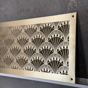 May include: A rectangular, gold-toned metal vent cover with an Art Deco fan pattern. The intricate design features repeating curved lines and geometric shapes. The vent cover is mounted on a dark gray wall.