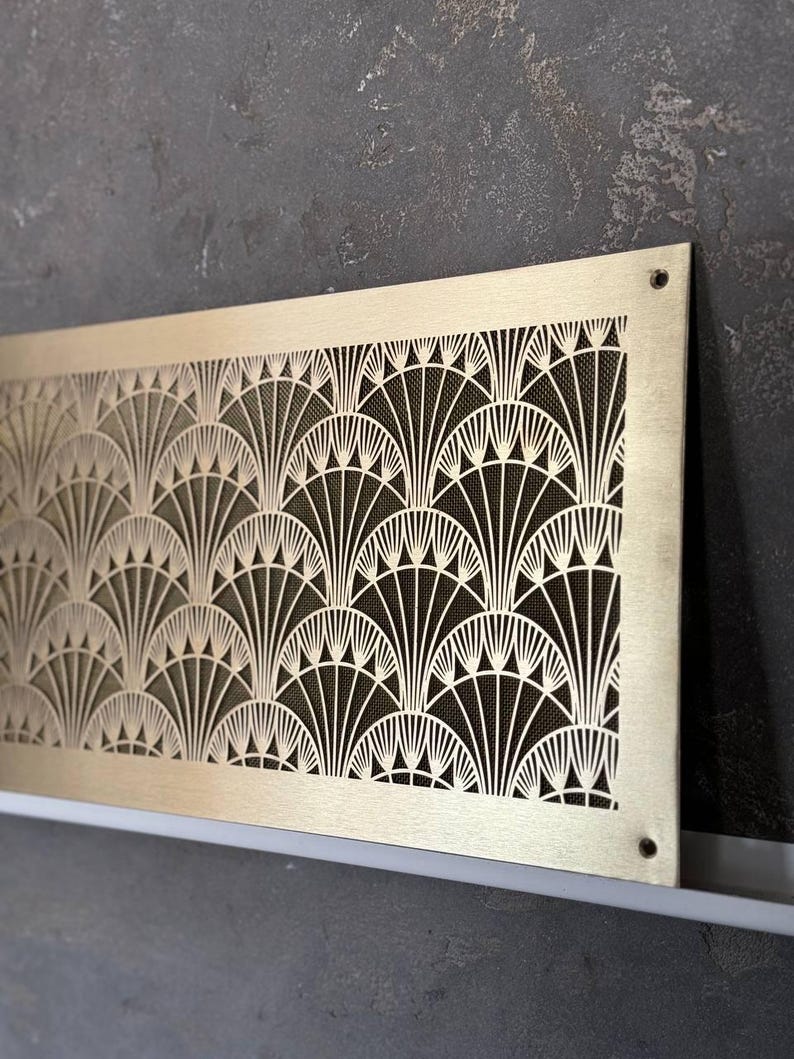 May include: A rectangular, gold-toned metal vent cover with an Art Deco fan pattern. The intricate design features repeating curved lines and geometric shapes. The vent cover is mounted on a dark gray wall.