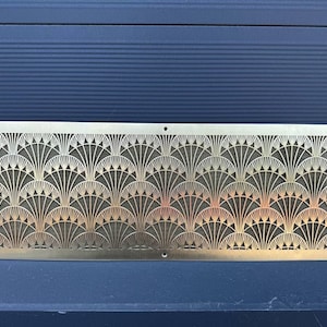 May include: A rectangular, gold-colored metal decorative panel with an Art Deco fan pattern. The panel has a series of repeating fan-shaped designs, with small holes at each corner and along the bottom edge.