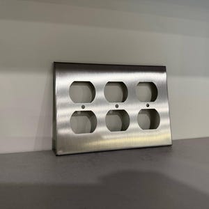 May include: A brushed stainless steel six-outlet wall plate. The plate has six openings for electrical outlets and is designed to cover and protect the electrical connections. The plate is rectangular and has a clean, modern design.