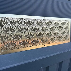 May include: A rectangular, Art Deco-style brass vent cover with a repeating fan-shaped pattern. The intricate design is cut out, revealing a dark mesh backing. The vent cover has a warm, metallic finish and is mounted on a dark surface.