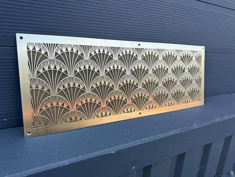 May include: A rectangular, Art Deco-style brass vent cover with a repeating fan-shaped pattern. The intricate design is cut out, revealing a dark mesh backing. The vent cover has a warm, metallic finish and is mounted on a dark surface.