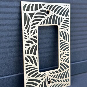 May include: A rectangular light wood switch plate cover with a leaf design cut out. The cover has a rectangular opening in the center and two holes for screws. The background is a dark blue surface.
