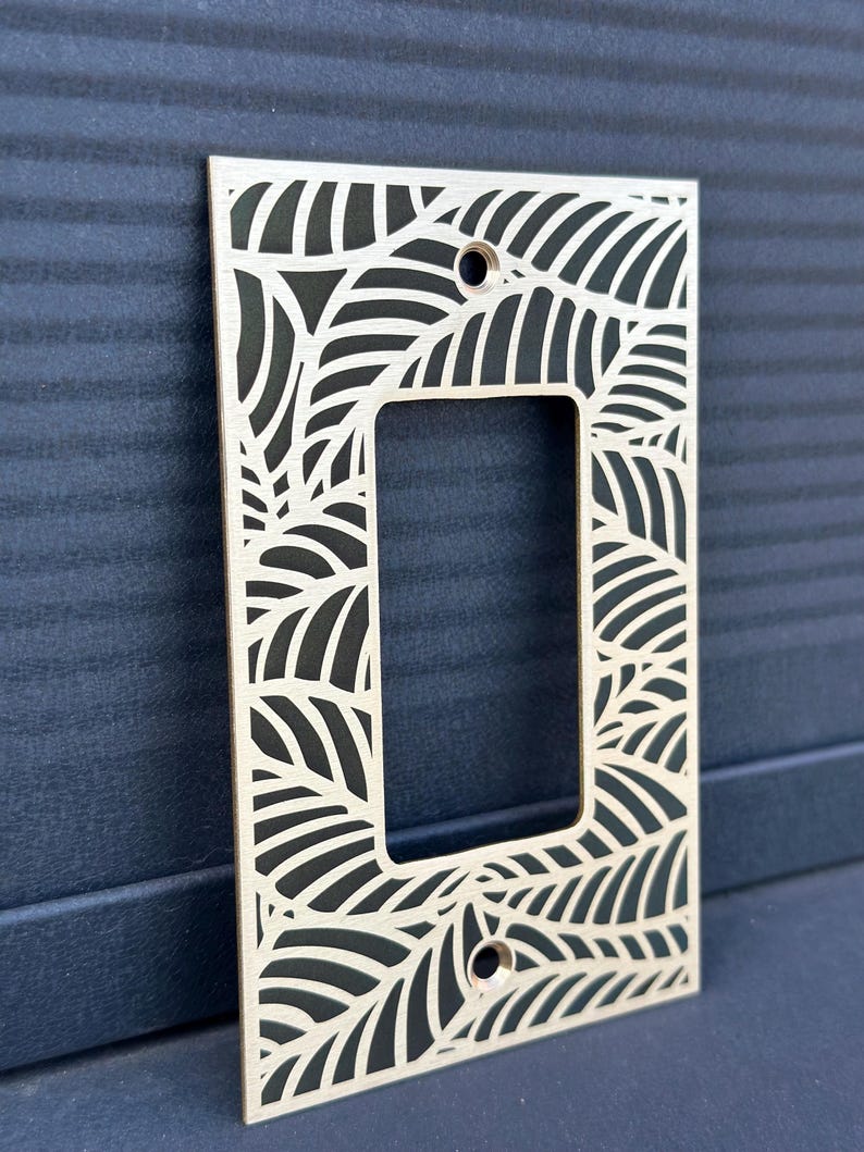 May include: A rectangular light wood switch plate cover with a leaf design cut out. The cover has a rectangular opening in the center and two holes for screws. The background is a dark blue surface.