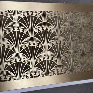May include: A rectangular, Art Deco-style brass vent cover with a repeating fan-shaped pattern. The intricate design features a mesh backing, creating a contrast between the solid brass frame and the openwork design. The vent cover is a decorative element.