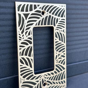 May include: A decorative light switch cover made of light-colored wood. The cover features a leaf design cut out around the edges and the center opening. Two holes are visible for mounting. The background is a dark blue.