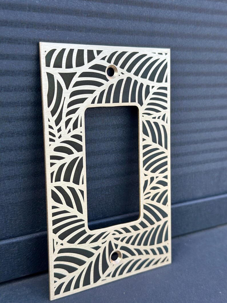 May include: A decorative light switch cover made of light-colored wood. The cover features a leaf design cut out around the edges and the center opening. Two holes are visible for mounting. The background is a dark blue.