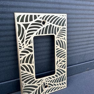 May include: A decorative light switch plate with a leaf pattern cut out of a gold-colored metal. The rectangular plate has a single opening for the switch and two screw holes. The leaf design is repeated across the surface.