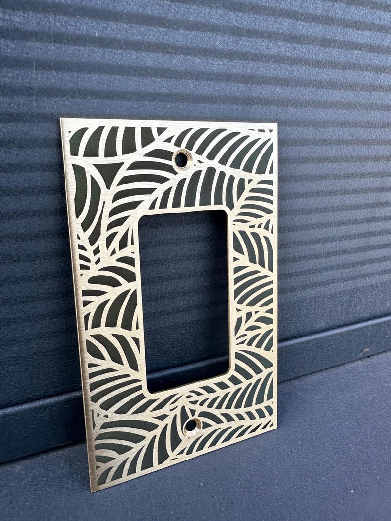 May include: A decorative light switch plate with a leaf pattern cut out of a gold-colored metal. The rectangular plate has a single opening for the switch and two screw holes. The leaf design is repeated across the surface.
