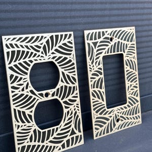 May include: Two decorative light switch covers with a leaf design. One cover has a dual outlet opening, and the other has a single switch opening. The covers are made of light-colored wood and feature a repeating leaf pattern.