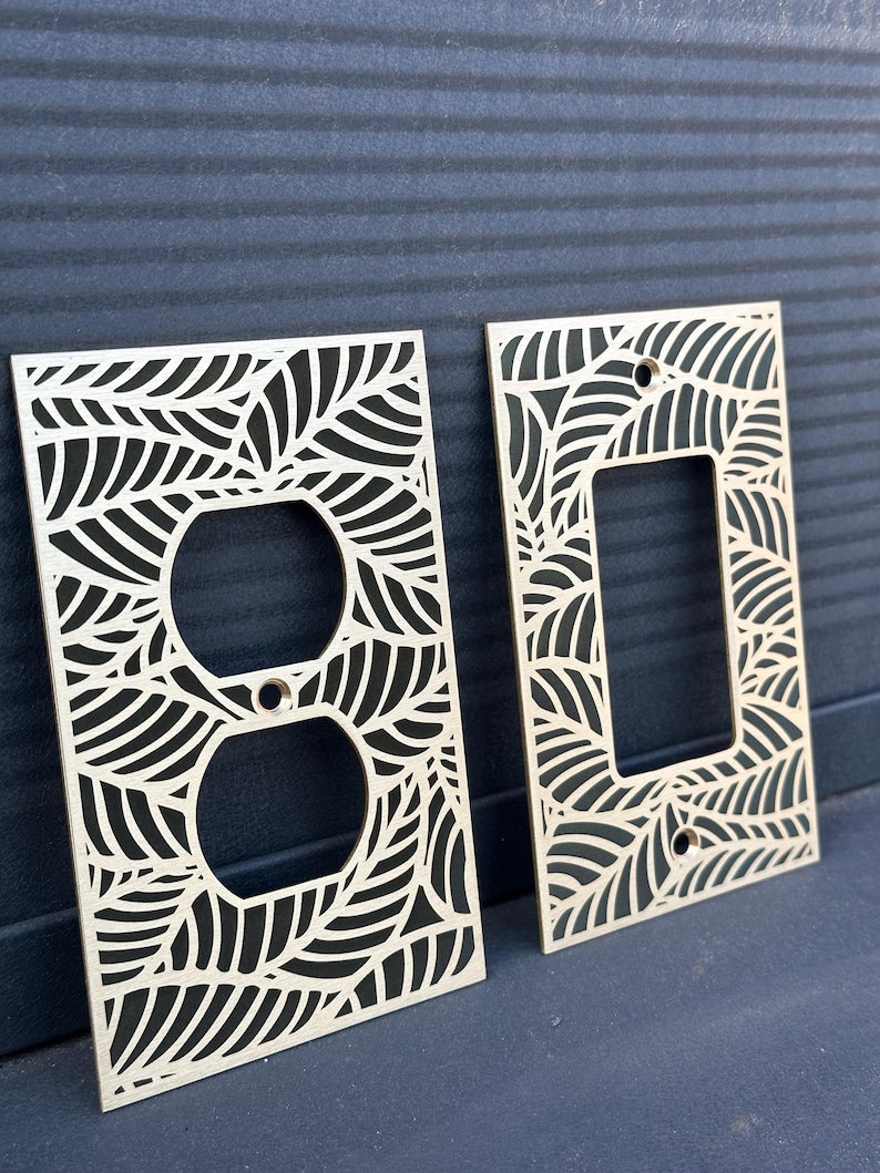 May include: Two decorative light switch covers with a leaf design. One cover has a dual outlet opening, and the other has a single switch opening. The covers are made of light-colored wood and feature a repeating leaf pattern.