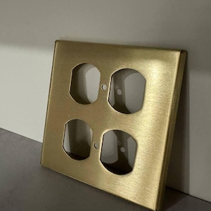 Könnte beinhalten: Eine quadratische, messingfarbene Steckdosenabdeckung mit vier Öffnungen. Die Abdeckung hat eine gebürstete Metalloberfläche und ist für Standard-Steckdosen geeignet. Die Abdeckung liegt auf einer grauen Oberfläche.