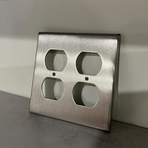 May include: A brushed stainless steel electrical outlet cover with four openings. The cover is rectangular with rounded corners and a smooth, metallic finish. The openings are oval-shaped, designed for standard electrical plugs.