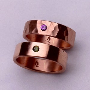 Op de afbeelding: Twee koperen ringen met gehammerde texturen. De bovenste ring heeft een paarse edelsteen en de onderste ring heeft een groene edelsteen. Beide ringen hebben de letter "Z" gegraveerd aan de binnenkant.