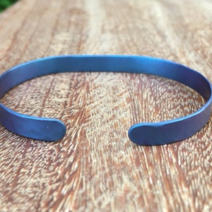 Op de afbeelding: Een blauwe titanium armband met een gehammerde afwerking. De armband is open en heeft een eenvoudig ontwerp.