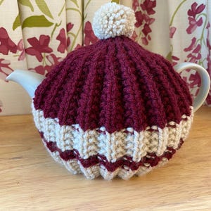 May include: A knitted tea cozy in burgundy and cream, topped with a cream-colored pom-pom. The cozy is designed to fit over a white teapot, with a ribbed pattern on the top and a decorative zig-zag pattern at the bottom.