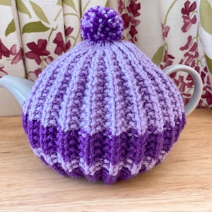 Pode incluir: Um aconchegante de chaleira de tricô roxo e lavanda com um pompom no topo. O aconchegante é feito com um padrão listrado.