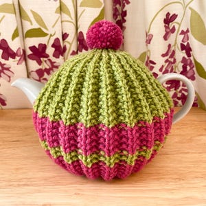 Può includere: Un copriteiera in maglia verde e rosa con un pompon in cima.
