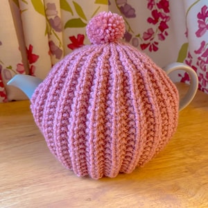 May include: A pink knitted teapot cosy with a pom-pom on top. The cosy is made with a ribbed stitch pattern.