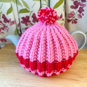May include: A knitted tea cosy in shades of pink and red, with a pom-pom detail. The cosy, with a ribbed design, sits on a light blue teapot. A decorative item for the kitchen, it is designed to keep tea warm.