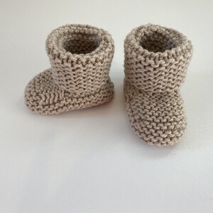 May include: A pair of beige knitted baby booties. The booties are made from a soft, textured yarn and have a simple, classic design.