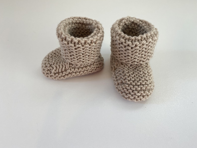 May include: A pair of beige knitted baby booties. The booties are made from a soft, textured yarn and have a simple, classic design.